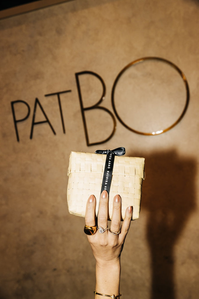 Hand holding a woven clutch with a 'PATBO' logo in the background