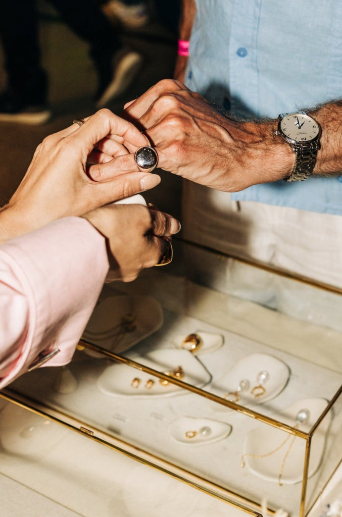 Two people holding hands over a jewelry box with rings inside, surrounded by people and decor.