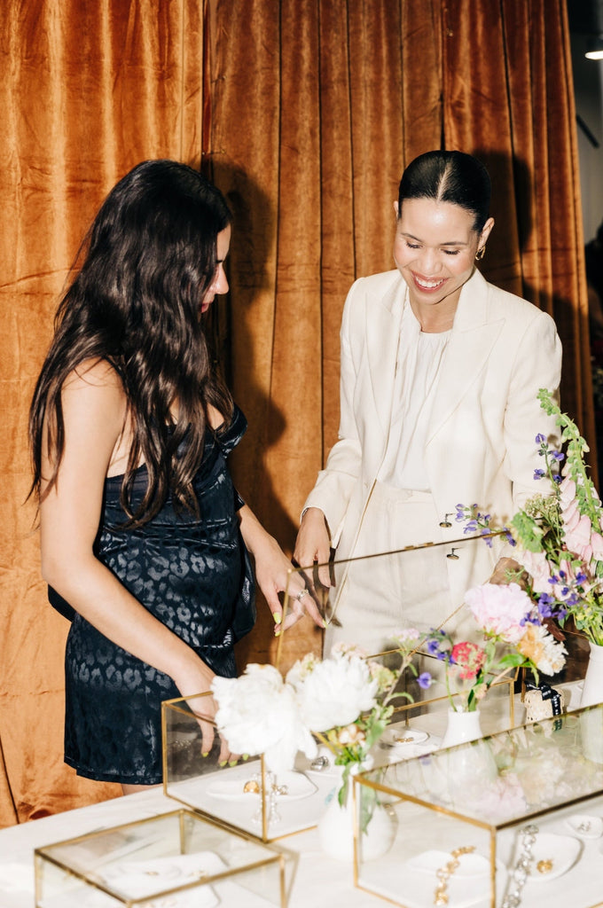 Two women standing at a table with floral arrangements and decorative items against a brown curtain.