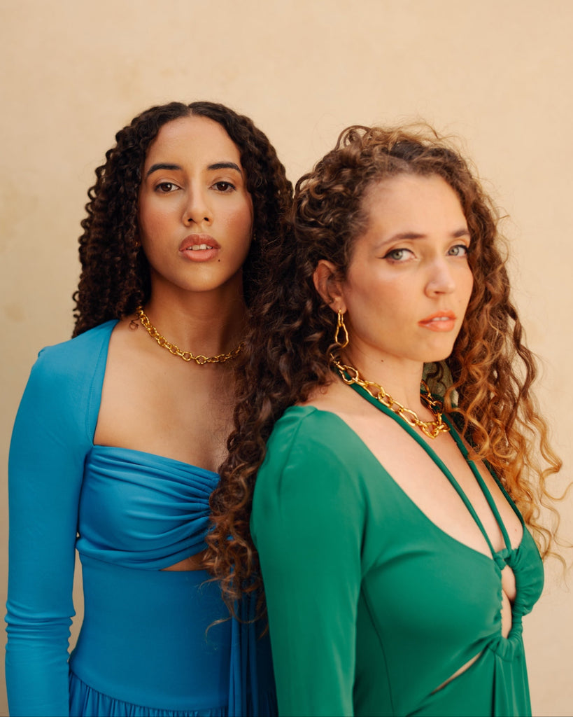 Two women standing back-to-back wearing blue and green dresses against a beige background